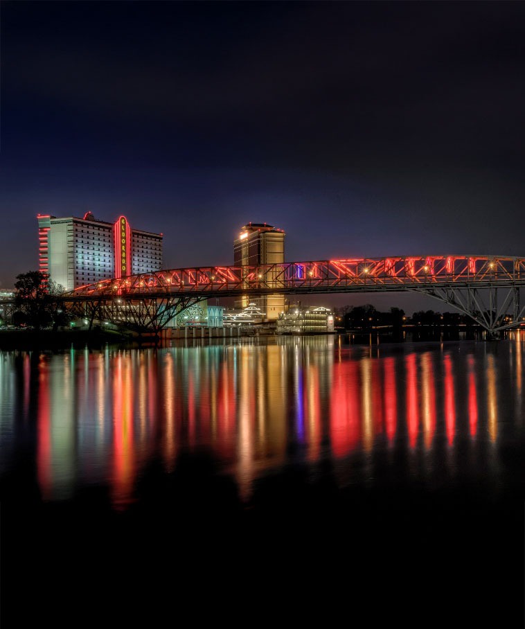 Nighttime cityscape with a brightly lit steel bridge spanning a calm river, neon red and yellow lights reflecting on the water, and illuminated high-rise buildings in the background.